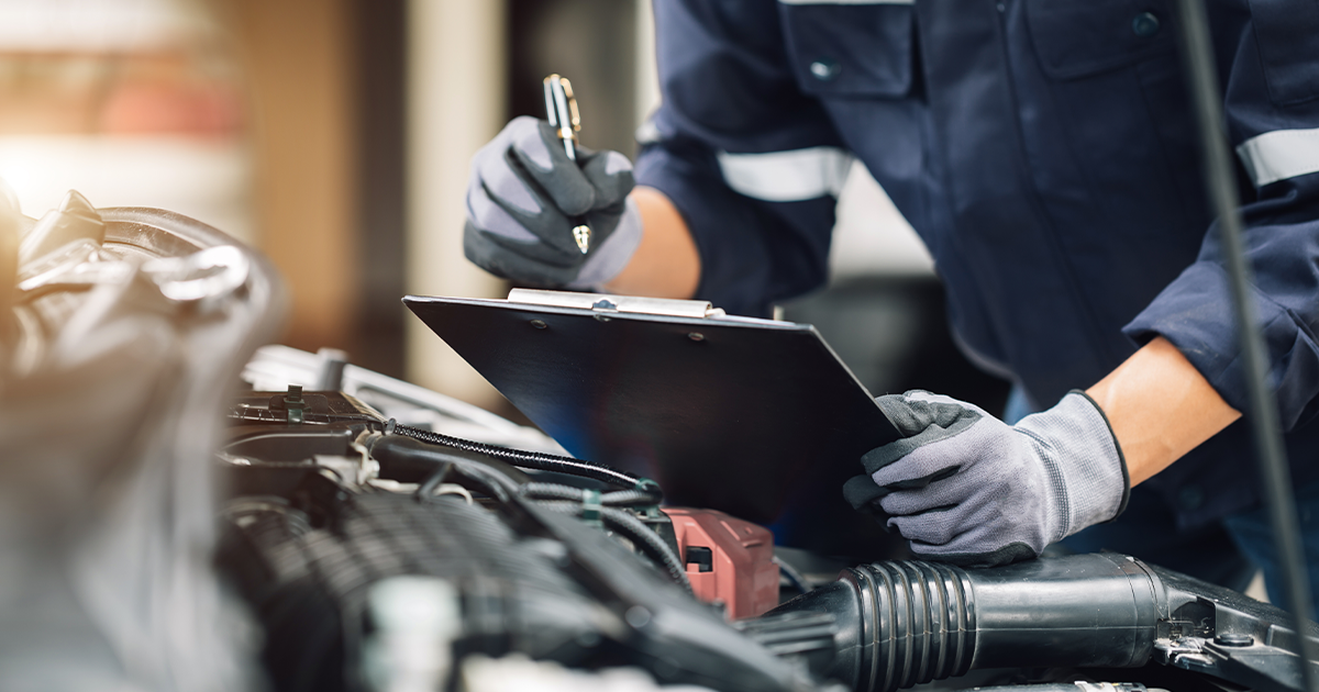 Close-up of a service technician holding a clipboard going through a checklist while inspecting a vehicle's engine.